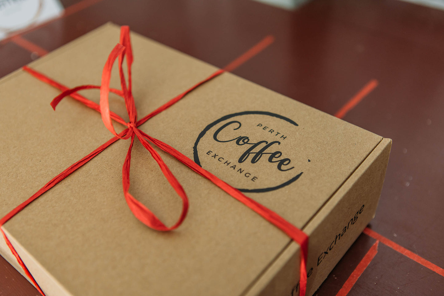 Perth gift-wrapped cardboard box with a red ribbon, labeled "Perth Coffee Exchange" in stylish black lettering, sits on a wooden table with red grains.