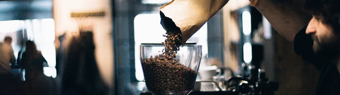 A person pours coffee beans from a paper bag into a glass container in a bustling cafe at Perth, capturing a moment of preparation in a warm, atmospheric setting.