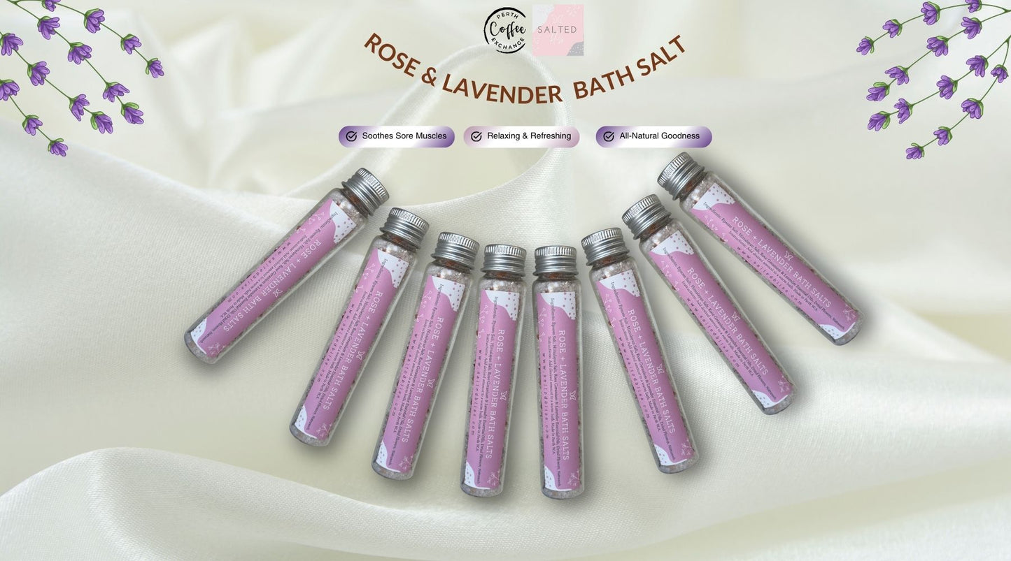 Image shows seven small glass vials of rose and lavender bath salts arranged on a silky white fabric surface, with lavender flowers suspended above. Each vial is labeled with benefits like soothing muscle aches and natural goodness in Perth.